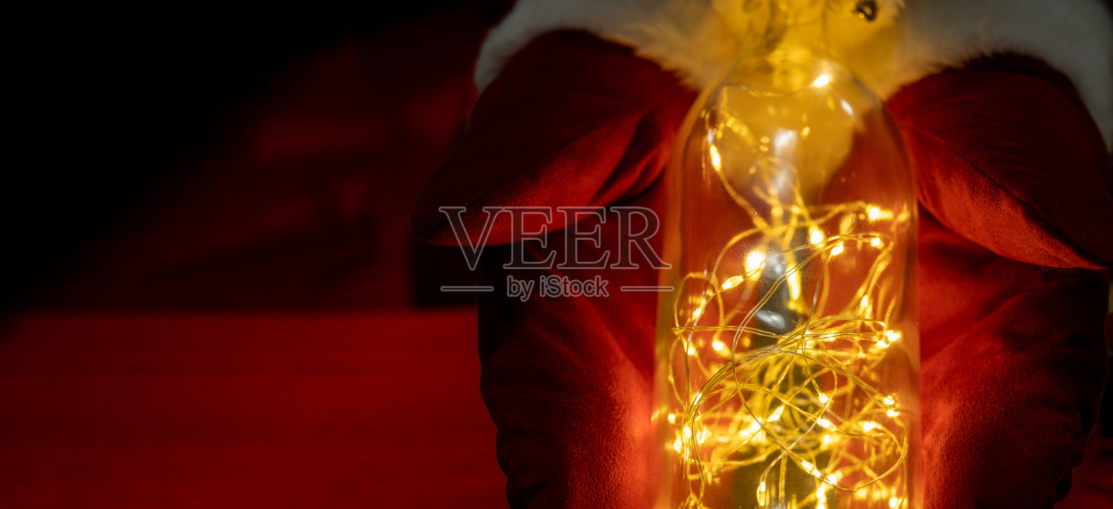 一个穿着圣诞老人衣服的人的手拿着一个有装饰灯的玻璃瓶。照片摄影图片