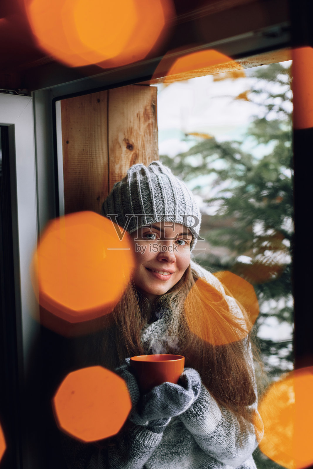 美丽的女人拿着并喝着一杯咖啡或可可在手套坐在家里的窗户旁。模糊的冬天雪树背景。早晨，舒适，冬天和人的概念照片摄影图片