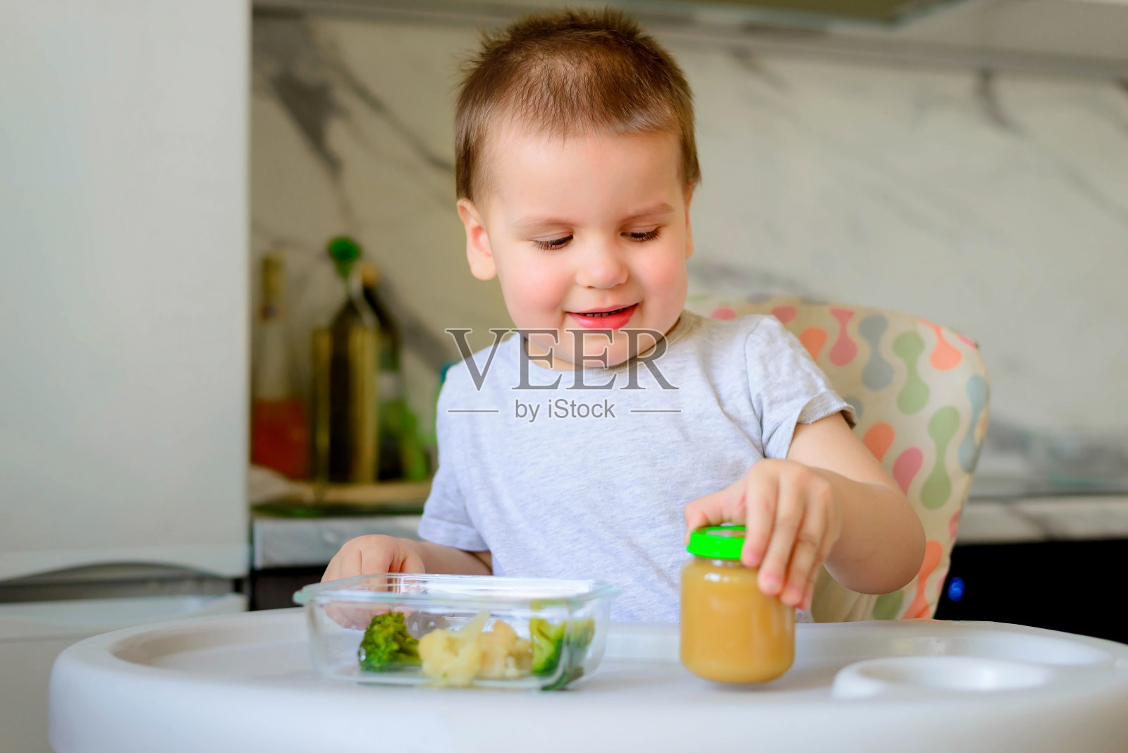 快乐的2岁男孩吃花椰菜，花椰菜和土豆泥在一个罐子在他的厨房在家里。婴儿食品，固体食品概念。照片摄影图片