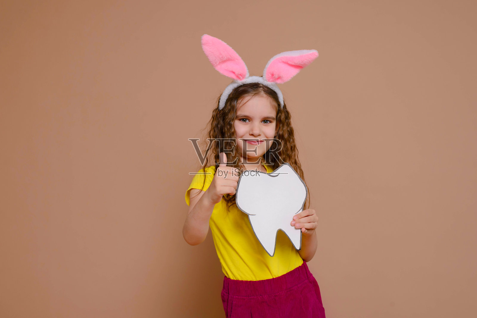 可爱的金发小女孩带着兔耳朵拿着一颗牙齿的模型。传统、打折、医药、庆祝的概念。照片摄影图片