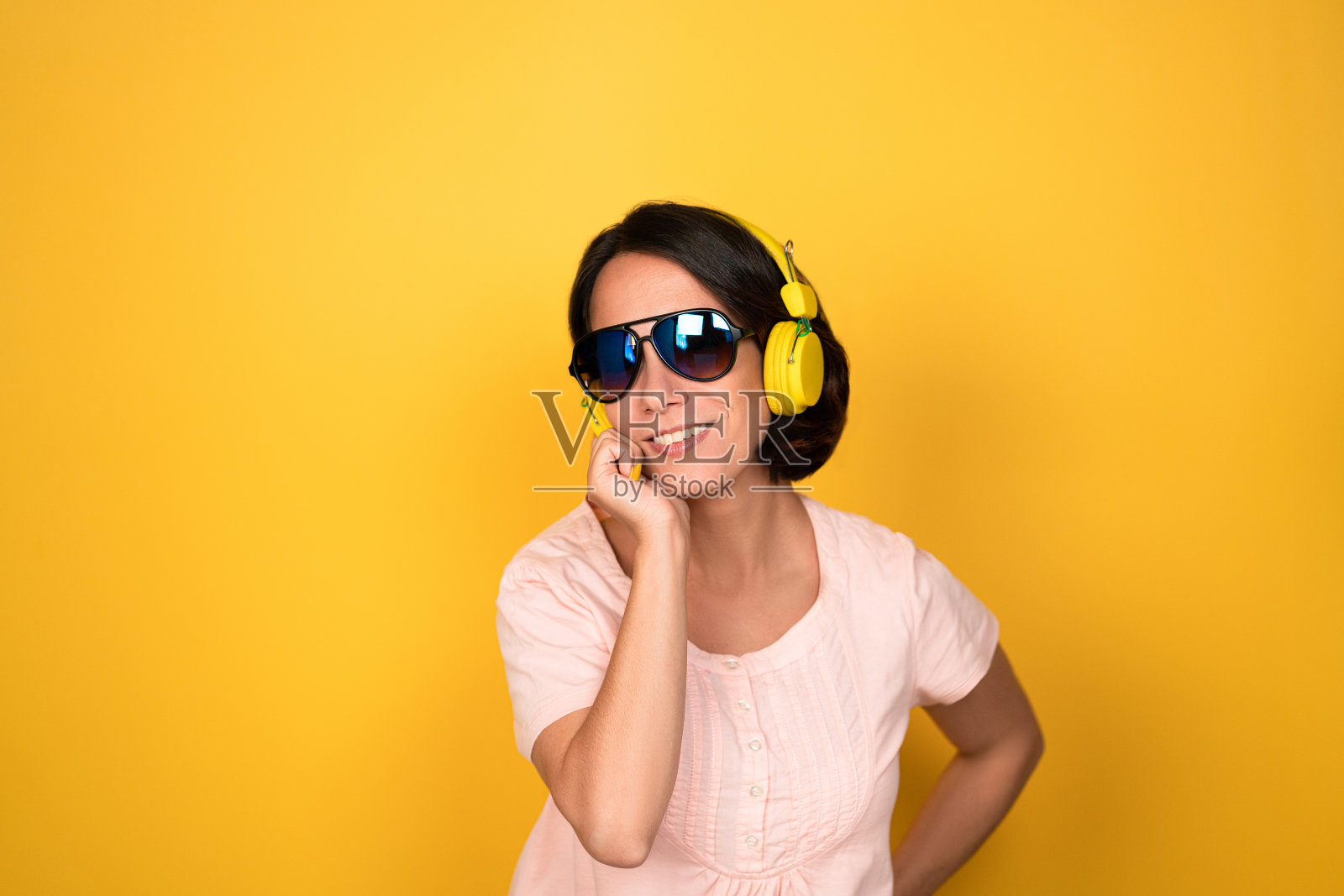 美丽的女孩的肖像在太阳镜和耳机听音乐，她触摸她的颧骨，牙齿的微笑，穿着休闲孤立的黄色背景照片摄影图片