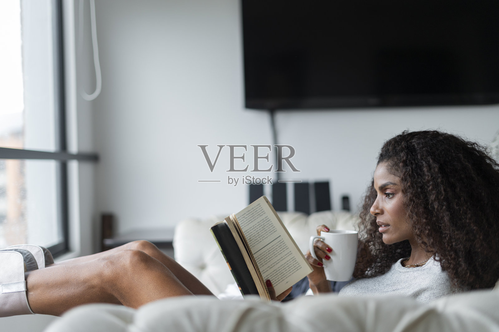 美丽的女人坐在她客厅的沙发上，旁边的窗户看她的书，喝着咖啡照片摄影图片