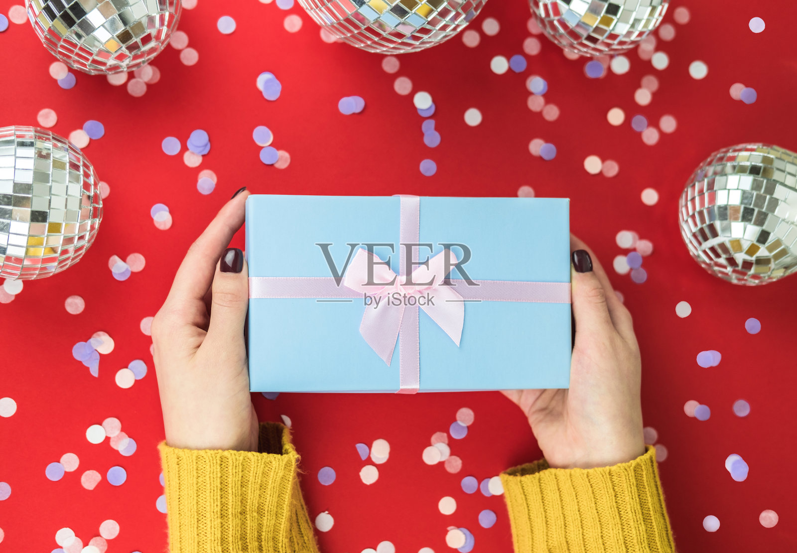 身穿橙色毛衣的女性手里拿着一个蓝色礼盒，盒子的背景是红色的，上面撒着五彩纸屑和迪斯科镜球。圣诞节和新年假期概念。从以上观点。平的。照片摄影图片