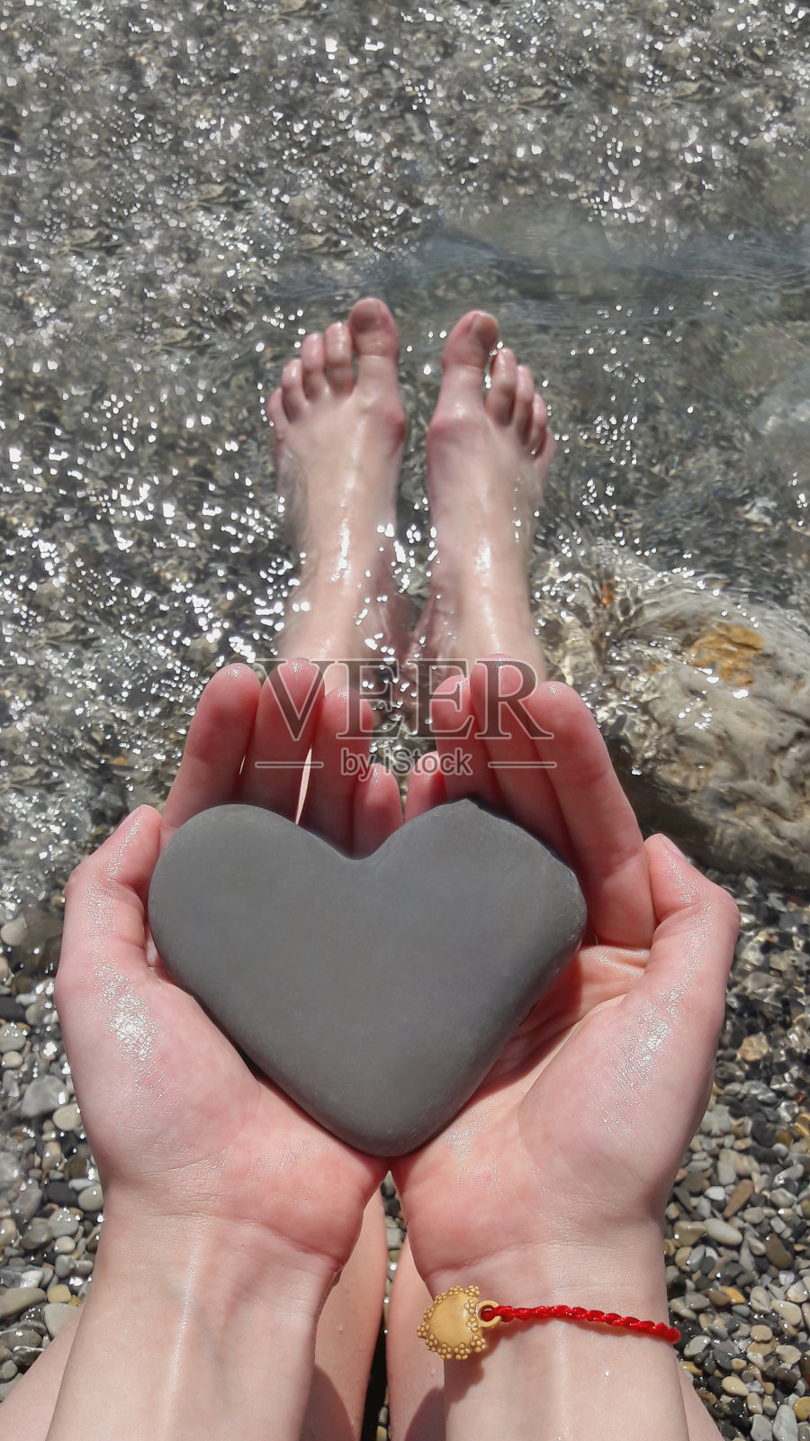 十几岁的女孩坐在卵石滩上的海，抱着一个天然的心形石头近距离。铁石心肠在手中。爱的概念。手机照片。照片摄影图片