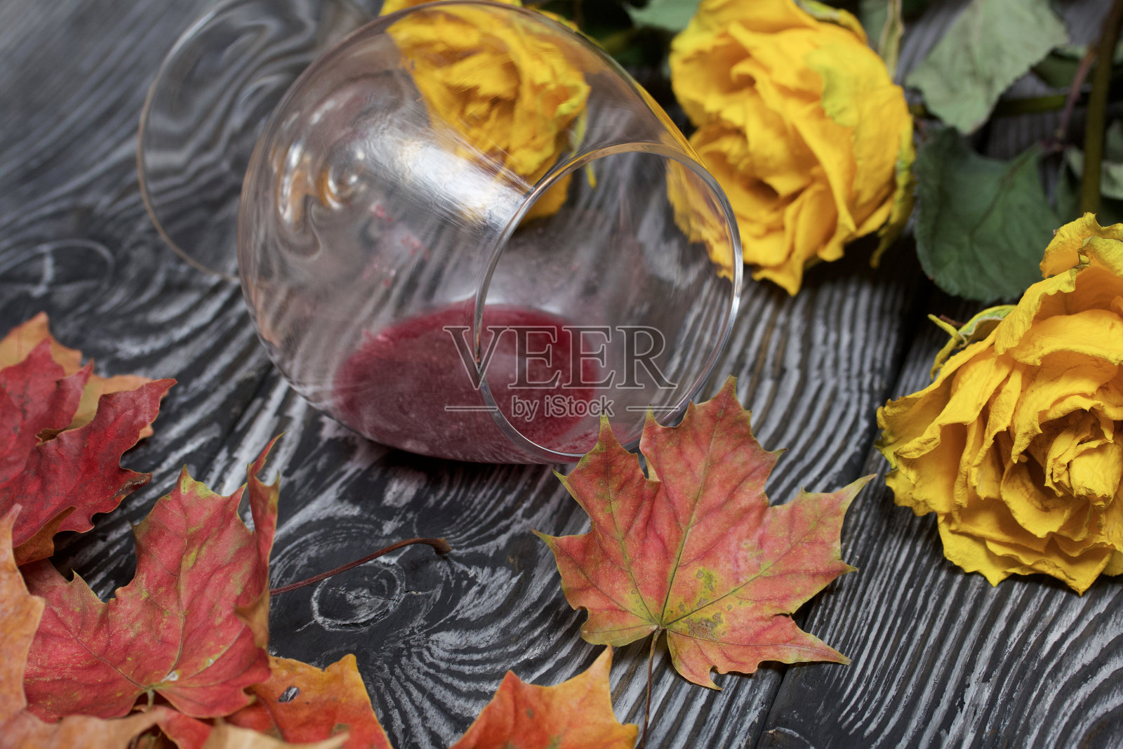 一束枯黄的玫瑰和干枯的秋叶。一杯打翻的酸酒。松木板。照片摄影图片