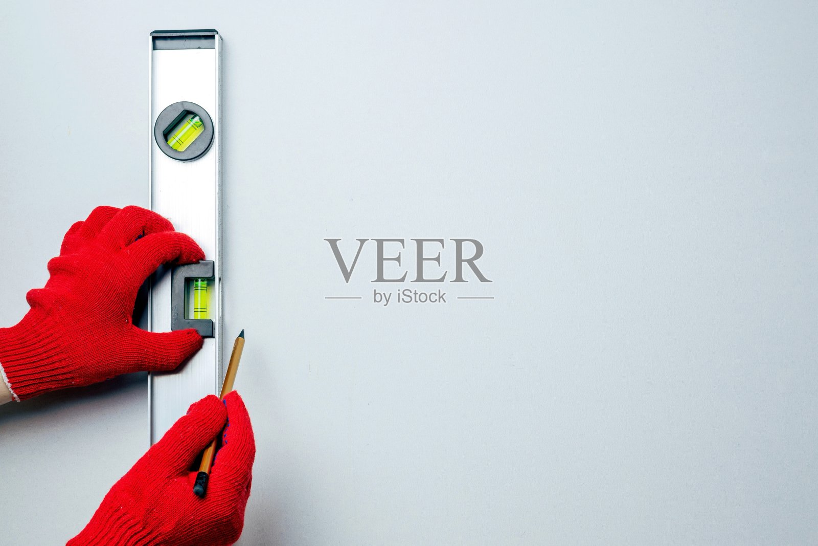 一个戴着红色防护手套的工人的手拿着一个金属水平仪和一支铅笔，靠在一堵白色的墙上。照片摄影图片