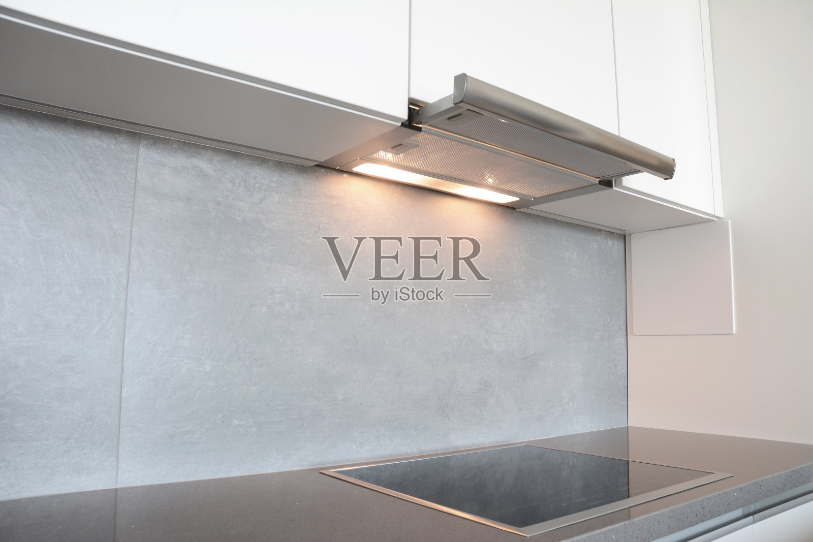 橱柜下的抽油烟机，带灯的排气罩，现代电炉，灰色和白色厨房设计的灶台。照片摄影图片