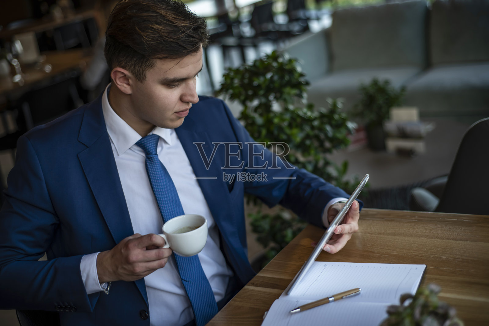 一个年轻的商人坐在咖啡馆里，计划着新的工作——一个穿着优雅西装的商人坐在窗边，一边思考，一边在他的平板电脑上工作照片摄影图片