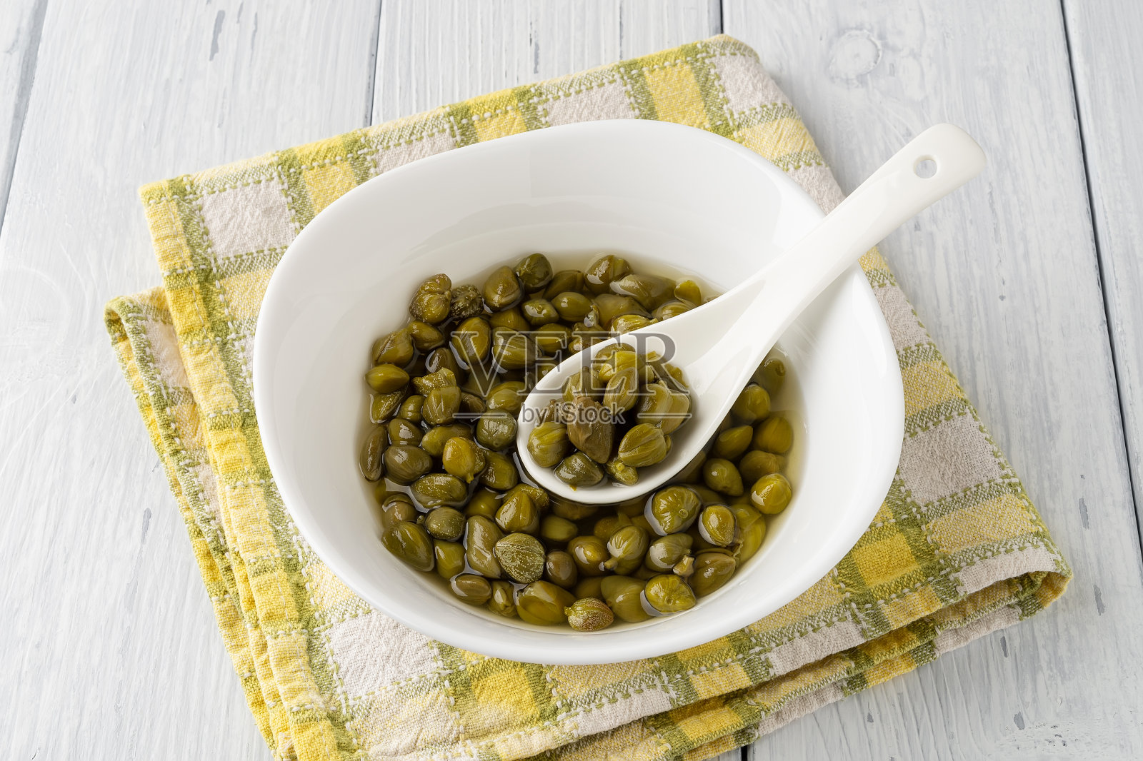 腌刺山柑放在盐水里，瓷勺放在白色木桌上的白碗里。腌过的刺山柑芽。地中海式饮食成分。有机香料和调味料。前视图。照片摄影图片