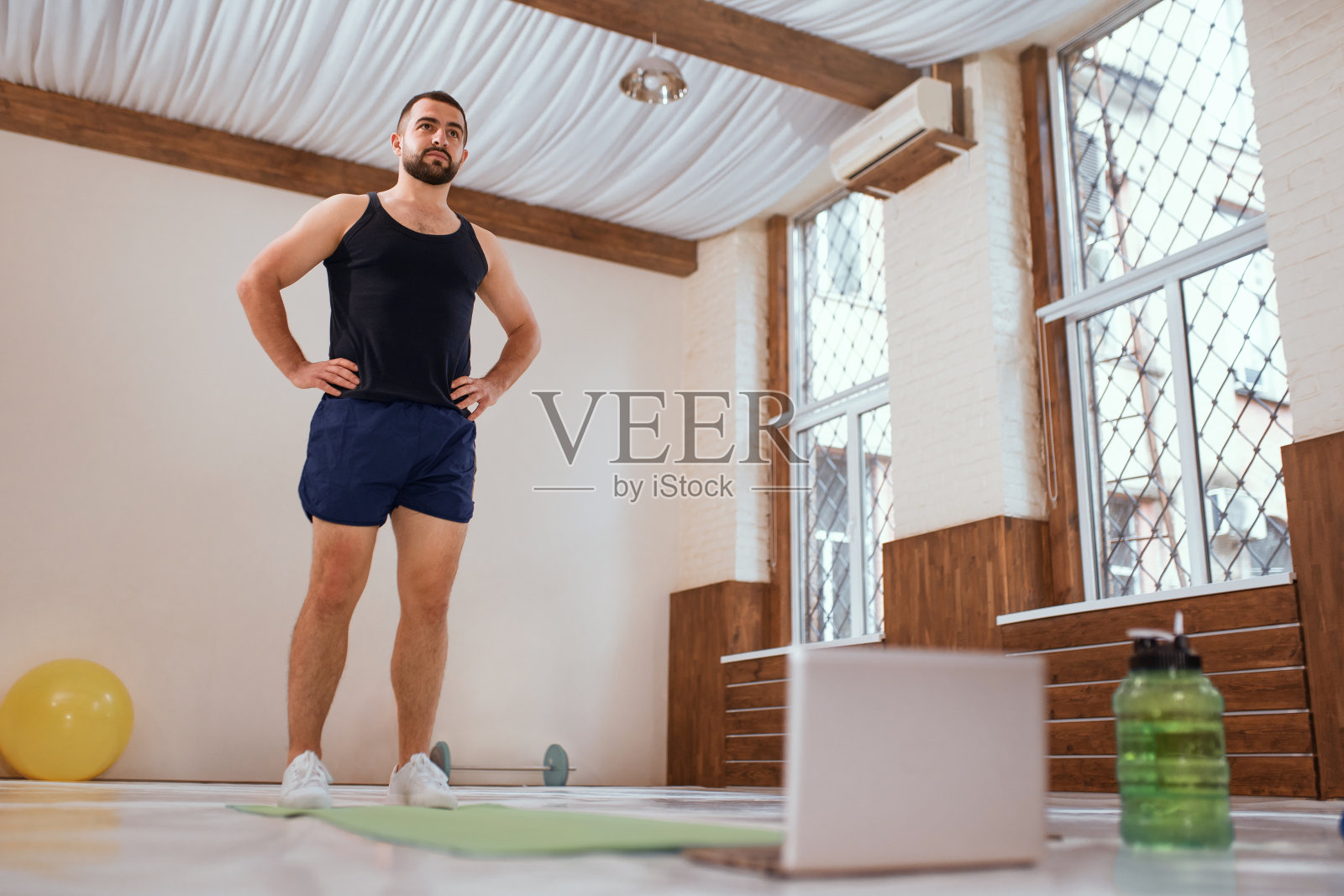 运动员在空荡荡的体育馆或家里站在笔记本电脑前刻苦训练。自我孤立和积极的年轻人热身做特殊的锻炼肌肉。在冠状病毒封锁期间工作照片摄影图片