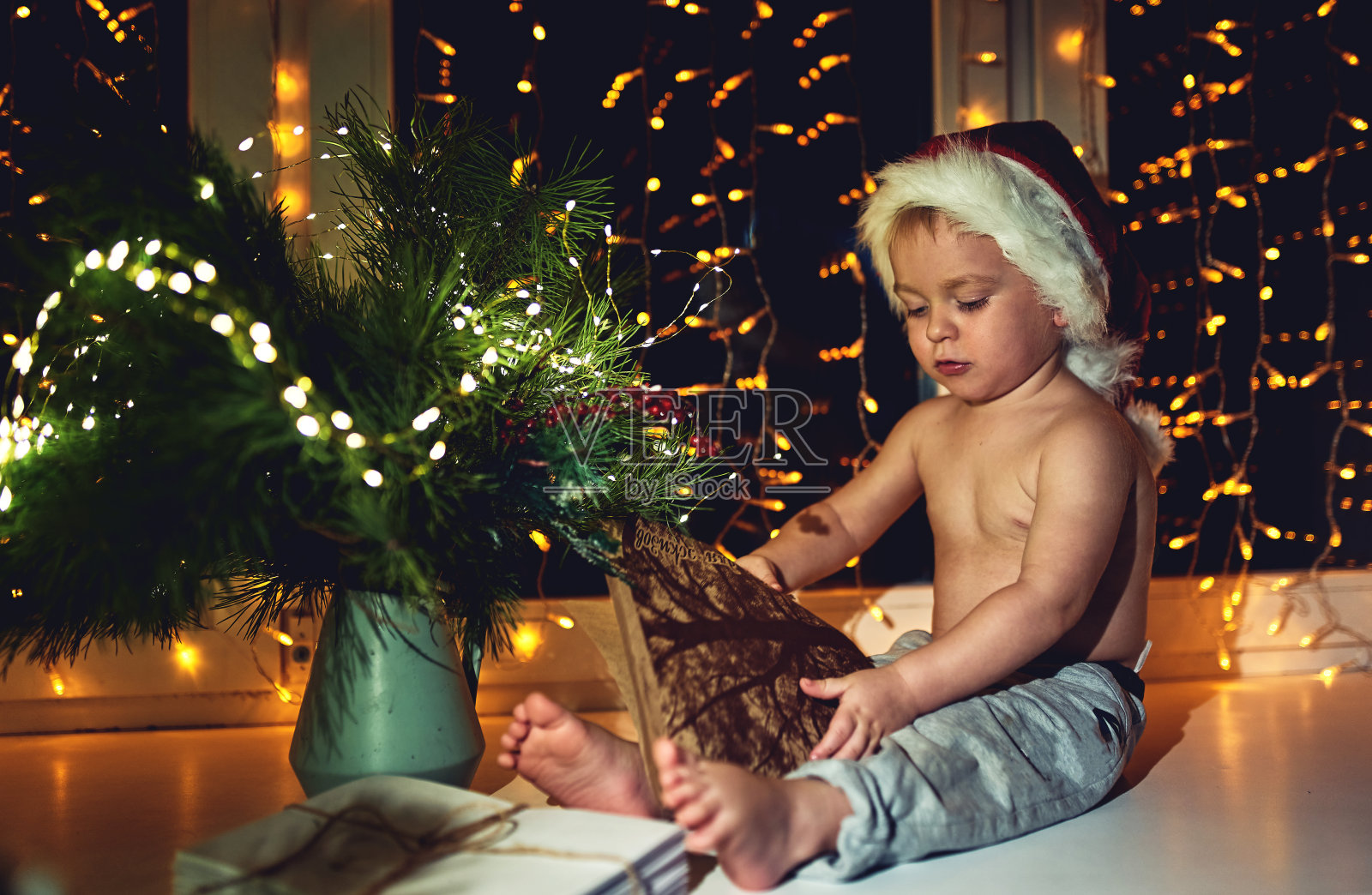 圣诞节的时候，一个戴着圣诞帽的漂亮小男孩坐在窗边照片摄影图片