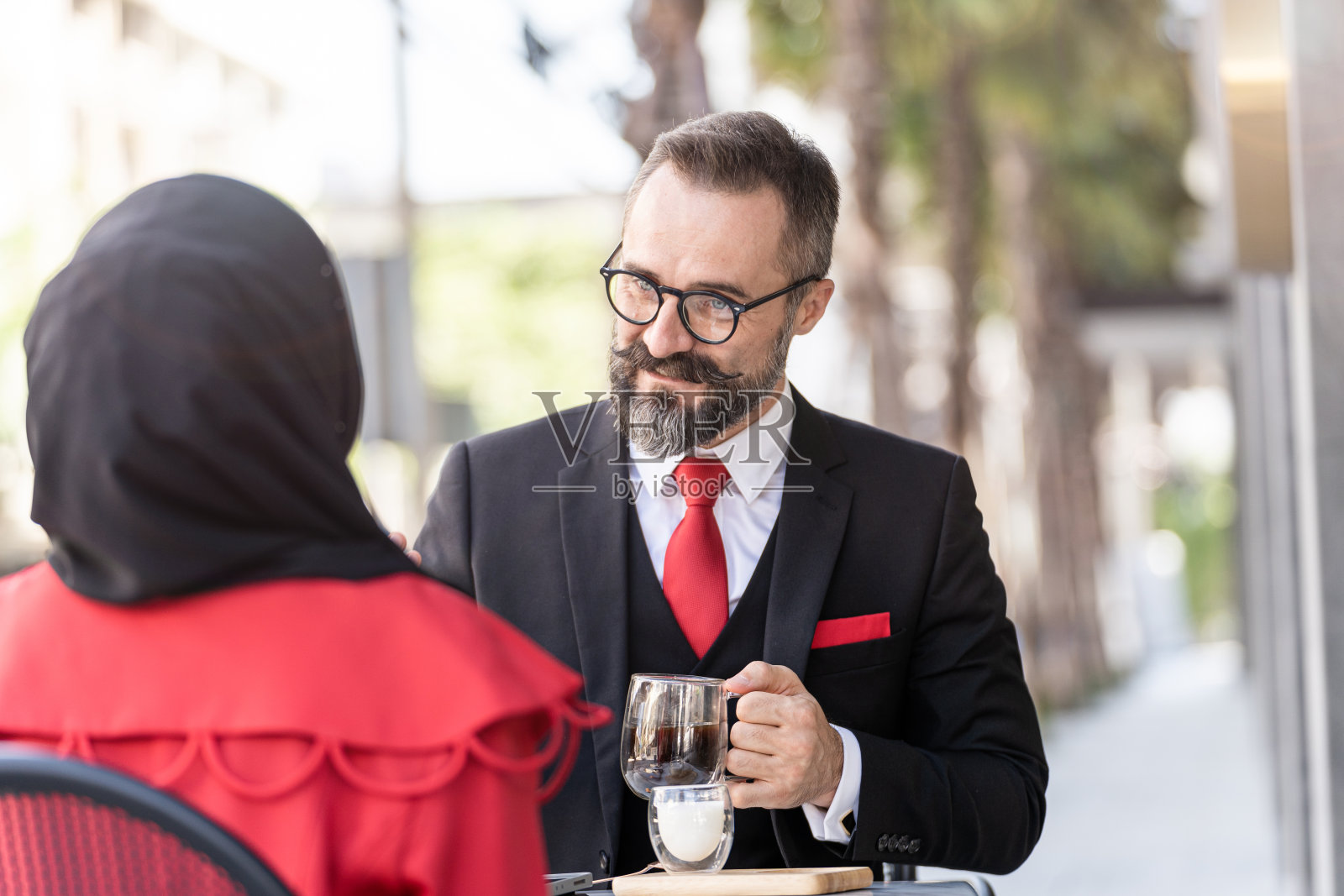一位穿着西装的高级男士坐在户外的咖啡桌上与人交谈。照片摄影图片