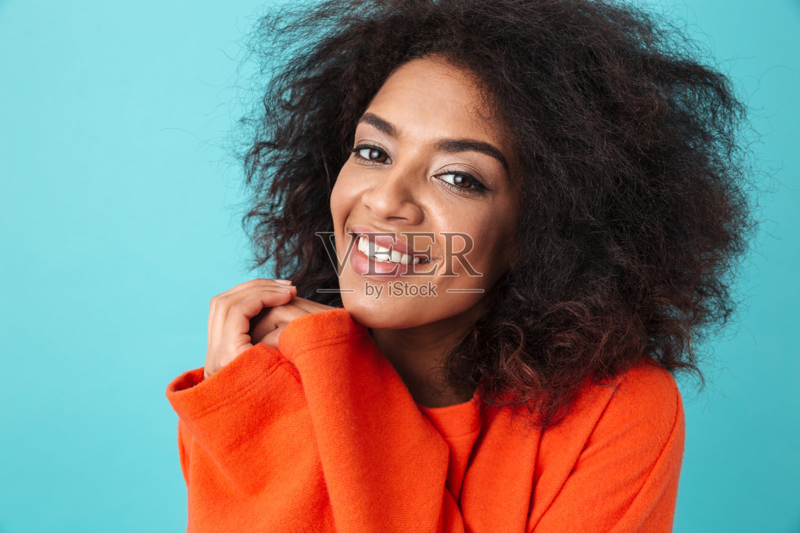 彩色肖像特写微笑的美国妇女在红色衬衫与非洲式发型看着镜头压她的手臂到胸部，孤立在蓝色的背景照片摄影图片