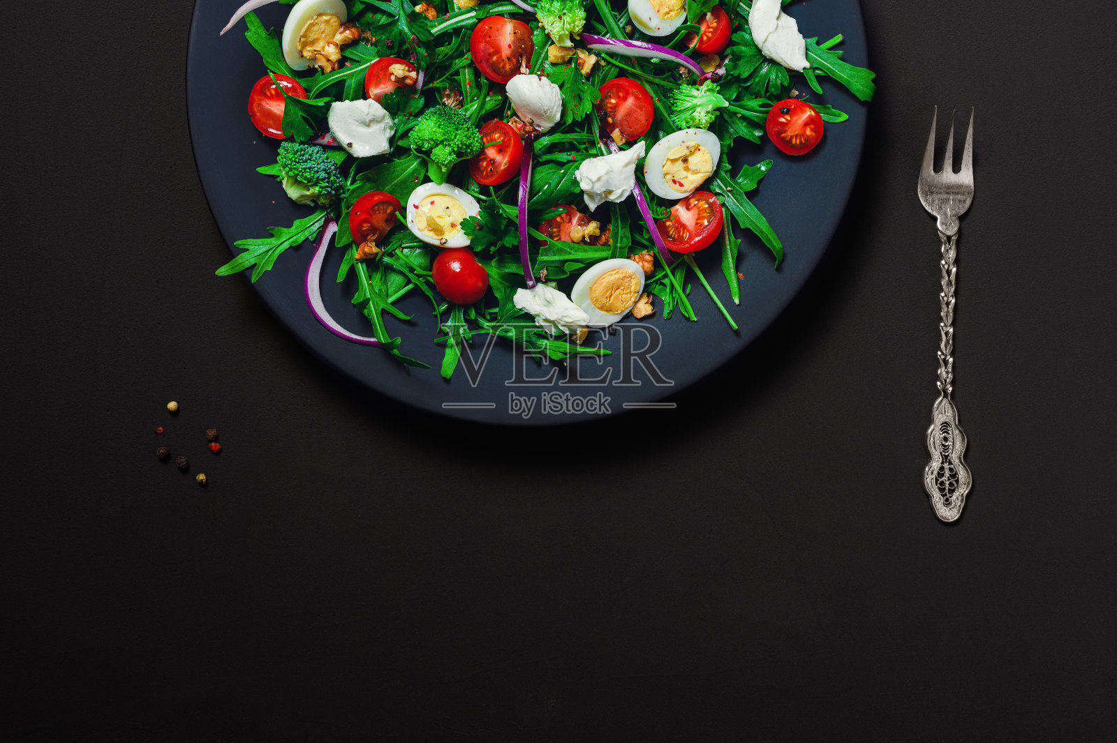 黑底黑底的深蓝色盘子，配上芝麻菜、西兰花、奶油芝士、樱桃番茄和核桃，用橄榄油调味。复制下方空间，平铺照片摄影图片