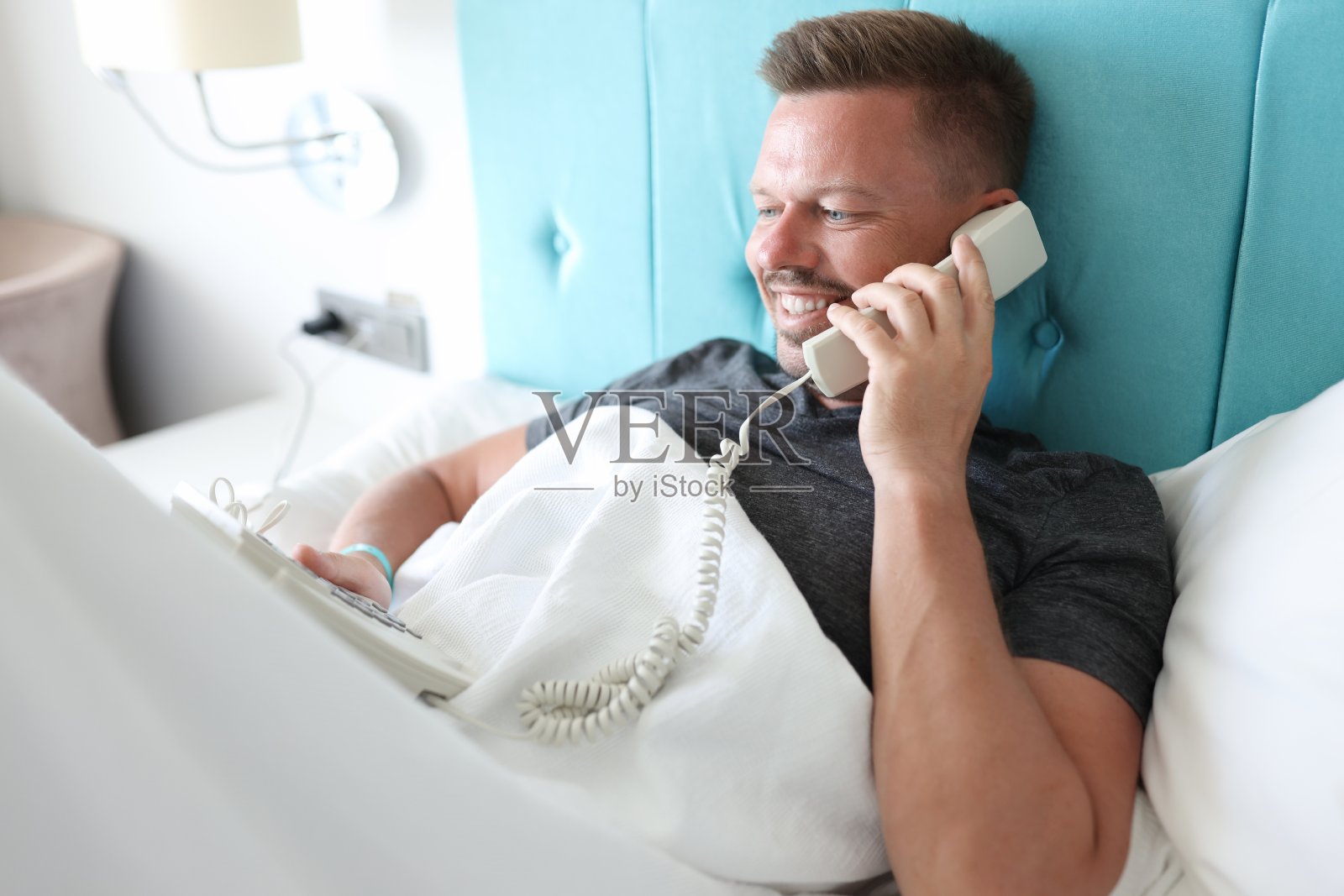 一名男子躺在酒店房间的床上，通过城市电话讲话。照片摄影图片