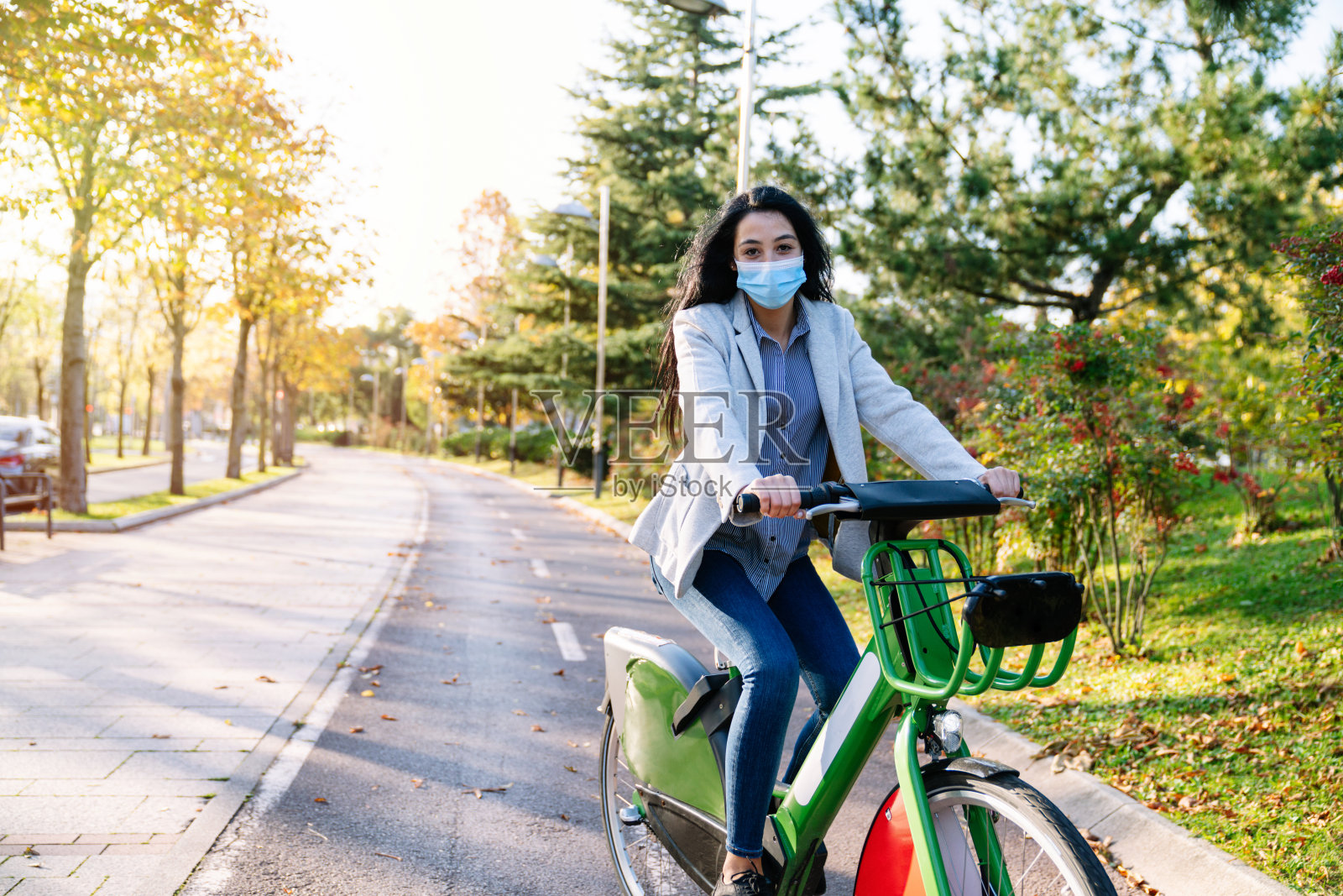 图为2020年2019冠状病毒(COVID-19)大流行期间，一名快乐工作的白人女孩骑着一辆共享电动自行车沿着林荫公园小路去上班，并戴着口罩照片摄影图片