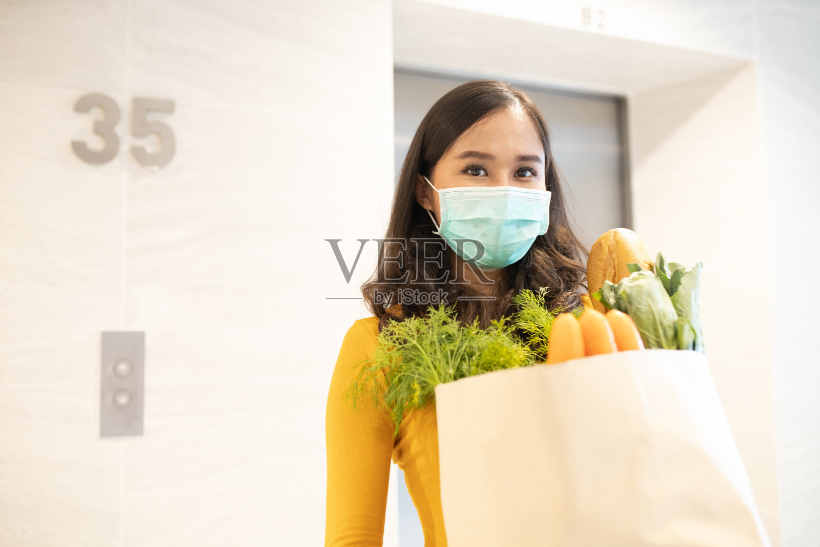 年轻的亚洲女性戴着医用口罩，手持一个生态纸袋，里面装着食品、水果和蔬菜、辣椒、长棍面包、生菜、安全在线配送、冠状病毒、检疫、大流行、居家理念。照片摄影图片