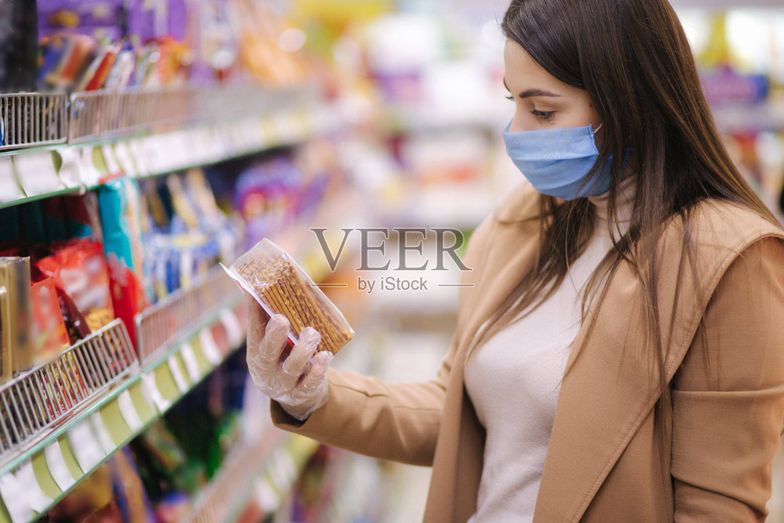 水平拍摄的妇女在保护口罩和手套阅读标签上的商品，而站在超市。美丽的年轻女孩在杂货店购物时选择蛇。在新冠肺炎大流行期间购物照片摄影图片