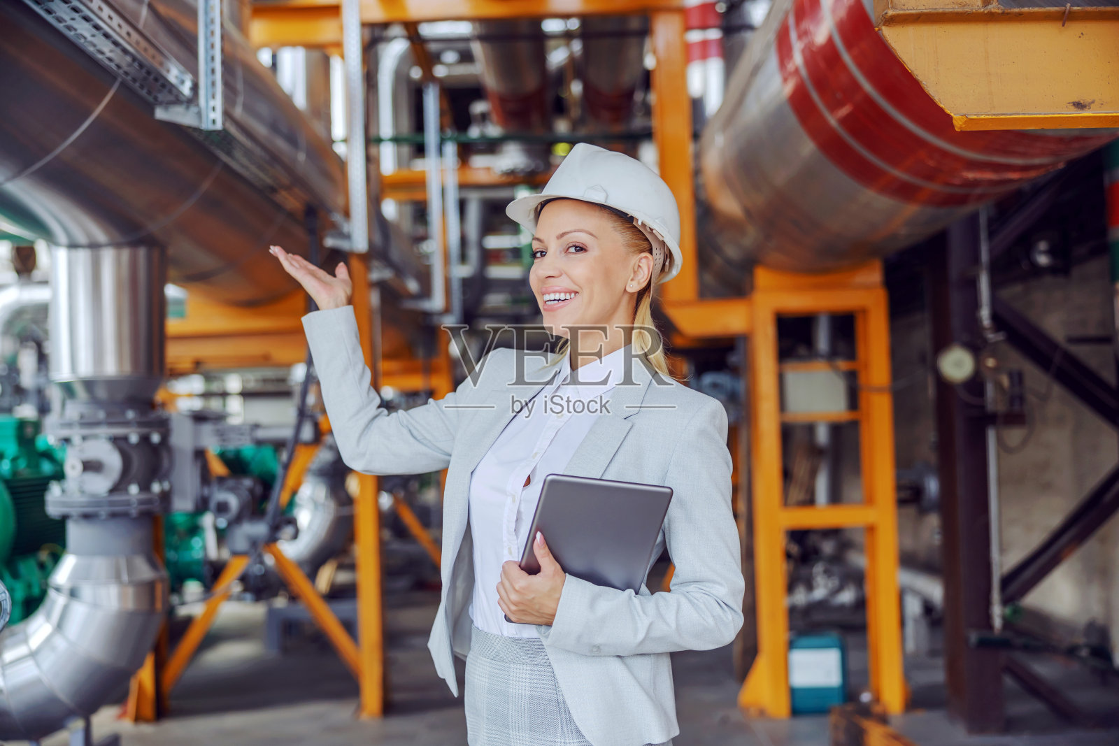 面带微笑的女CEO穿着正装，头戴防护头盔，手持平板，站在供热厂。照片摄影图片