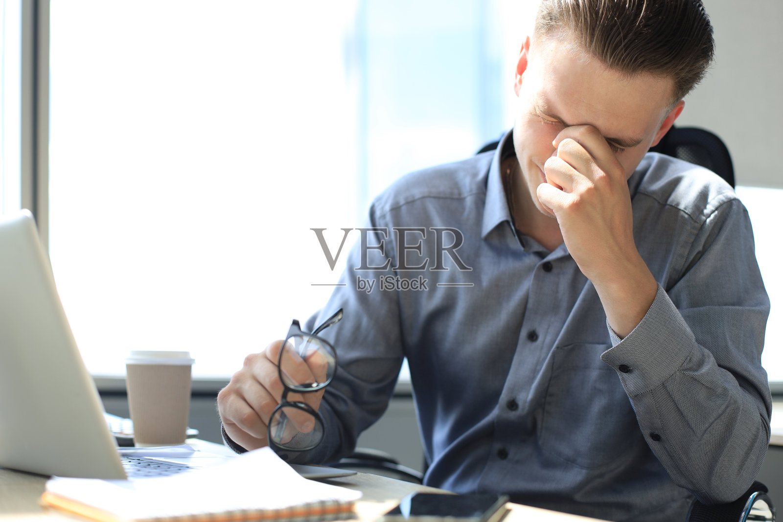 英俊的年轻商人闭着眼睛用手触摸脸，而坐在办公室工作的地方。照片摄影图片