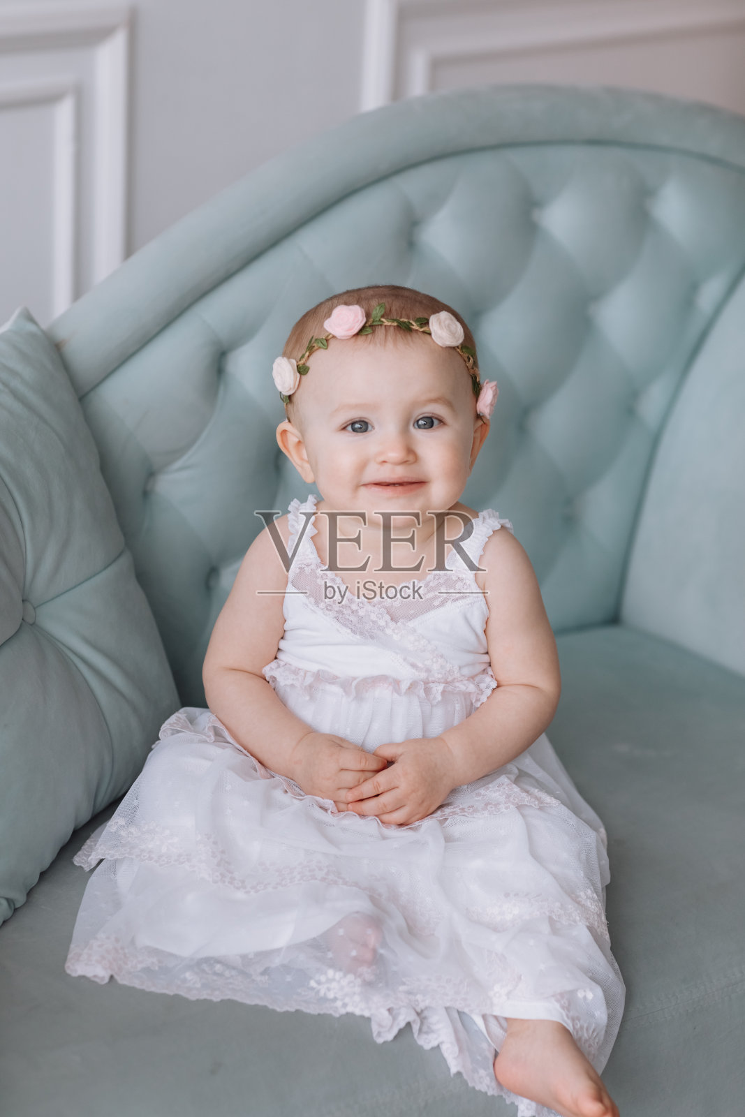 可爱的小女孩穿着白色的裙子，头上戴着花环，坐在沙发上，在家里玩得很开心。照片摄影图片