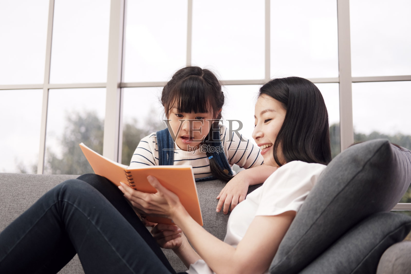微笑的亚洲母亲和小女孩在客厅的沙发上一起看书。家庭教育与教育理念照片摄影图片