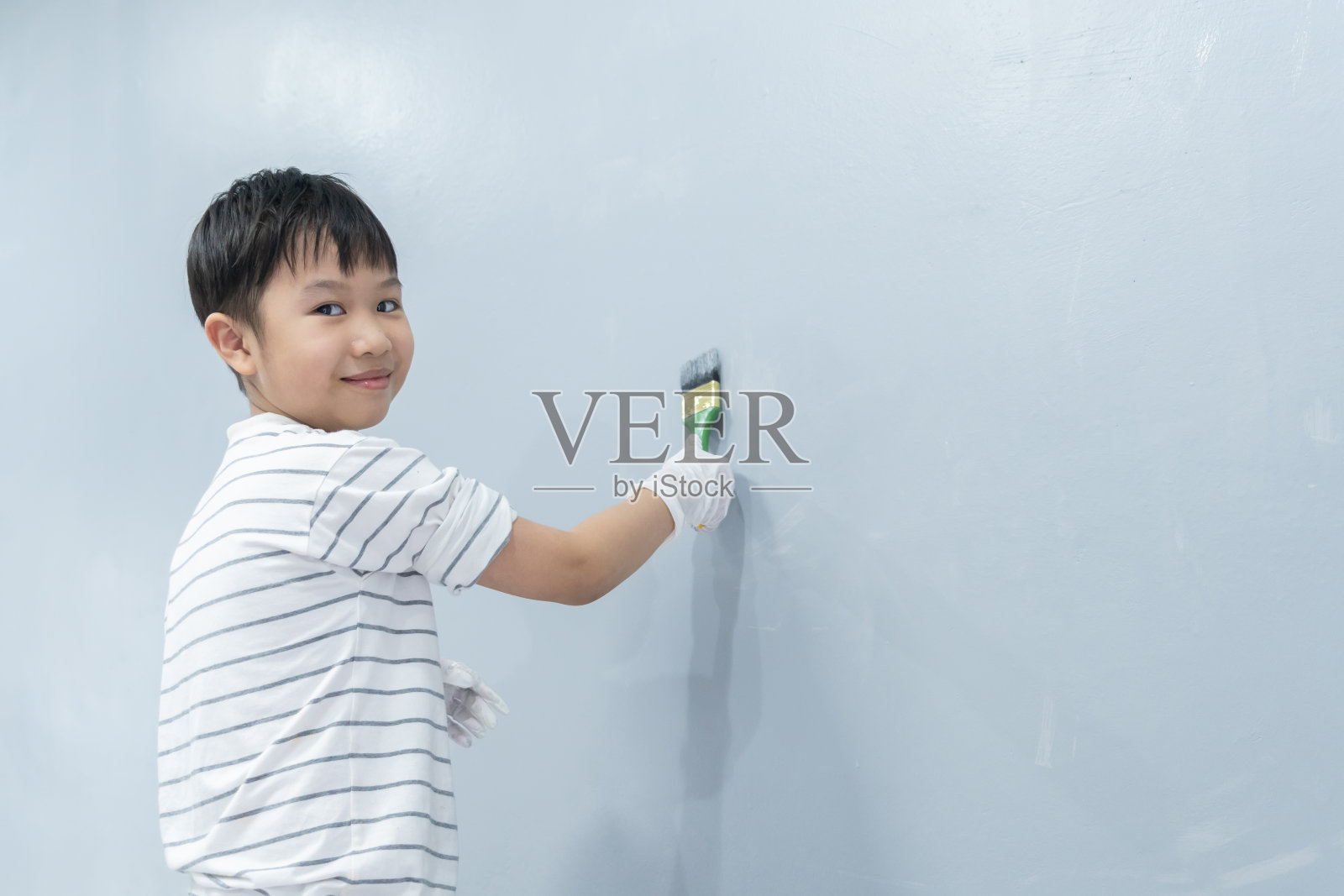 一个亚洲男孩用油漆滚筒把白墙漆成灰色。孩子们通过帮助做家务来练习技能。家居保养和装修理念照片摄影图片