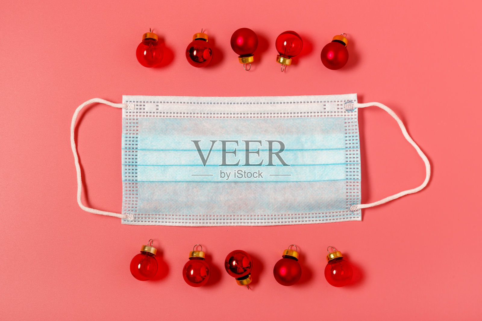红色圣诞球和红色背景上的蓝色外科口罩。冠状病毒大流行和疾病防护的概念。圣诞节假期的隔离概念。俯视图，平铺，最小。照片摄影图片