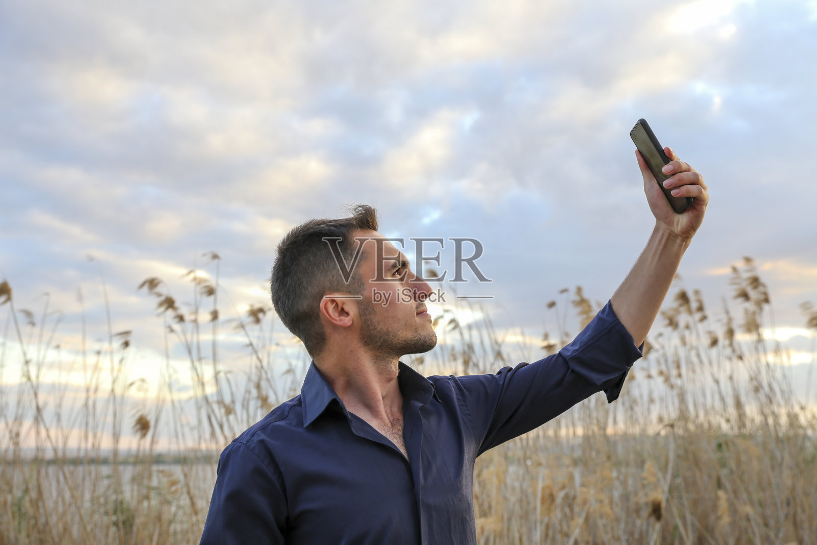 分享快乐的有问题。英俊的年轻微笑的男人穿着休闲装，用他的智能手机自拍，而站在户外。照片摄影图片