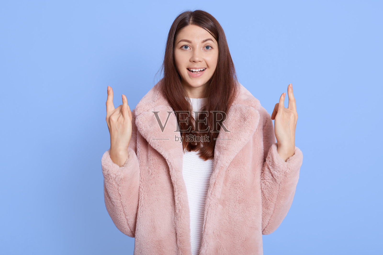 照片时髦可爱的年轻女子穿着淡粉色毛皮大衣显示硬岩石标志孤立在蓝色背景，看着相机与兴奋的面部表情。照片摄影图片