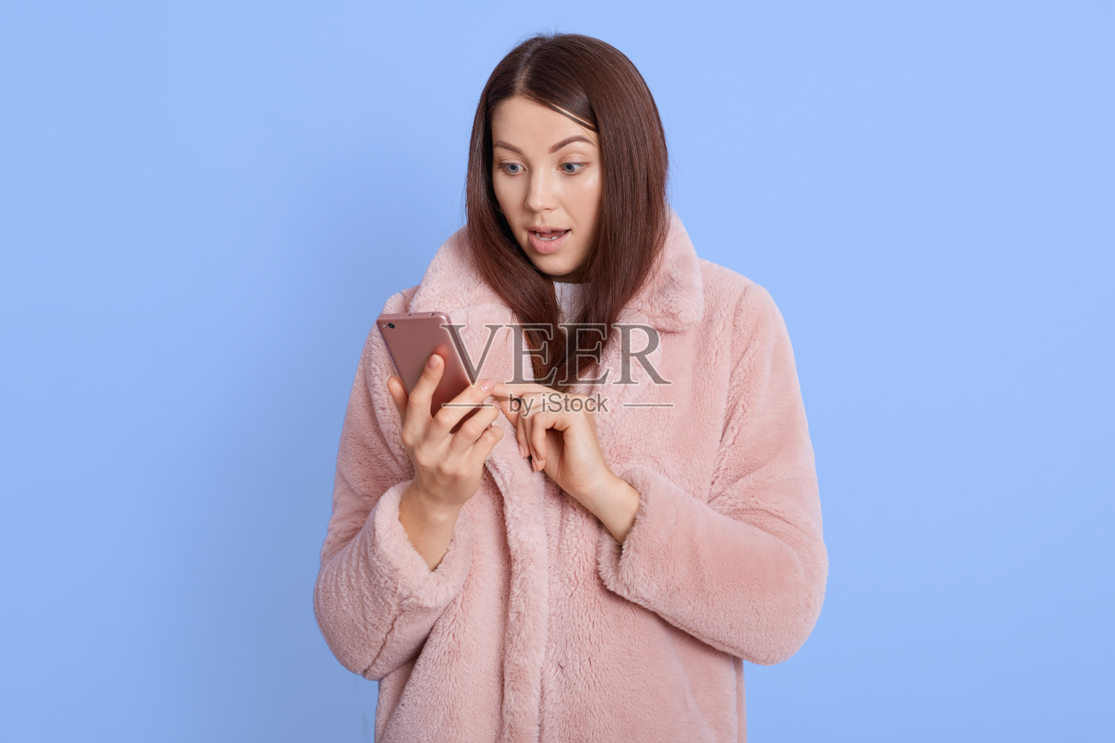 照片惊讶漂亮的女士拿着电话在手里，分享新鲜的新闻，穿着风格粉红色人造皮草大衣，震惊女性摆出孤立的蓝色背景。照片摄影图片