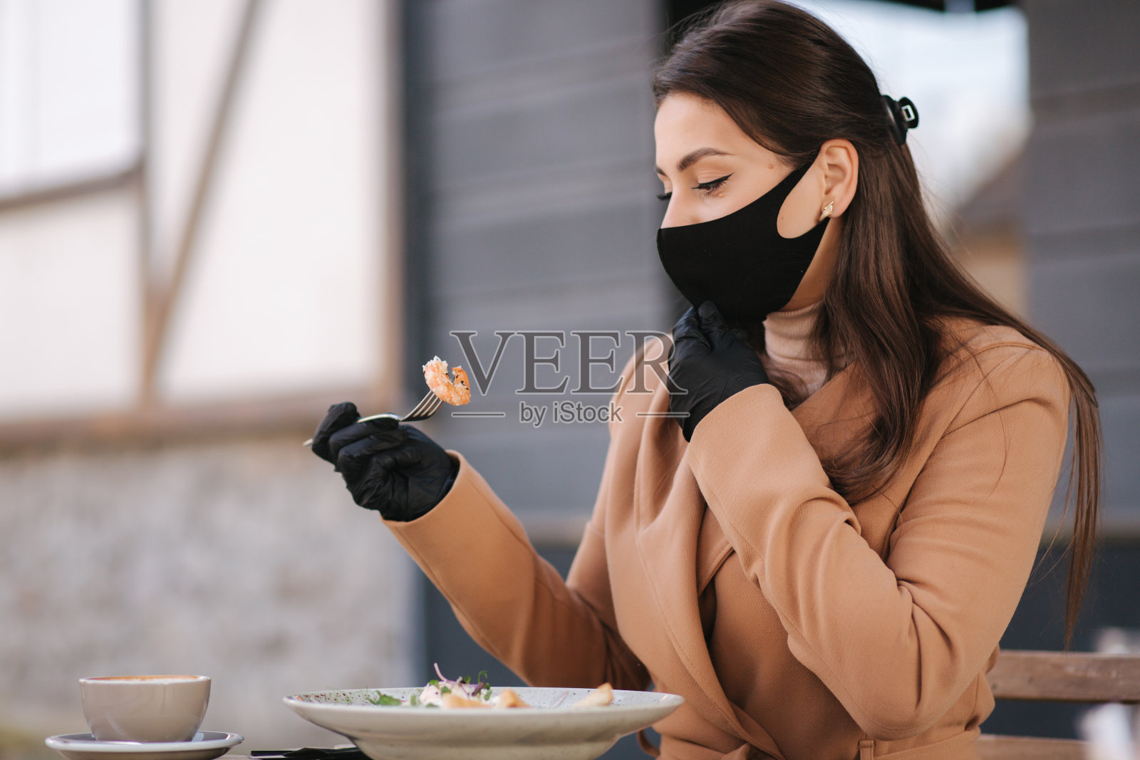 戴黑手套的女人拿着餐具。检疫咖啡馆的概念。带防护手套的户外食品。新鲜的素食虾沙拉。女性脱下防护面具进食照片摄影图片