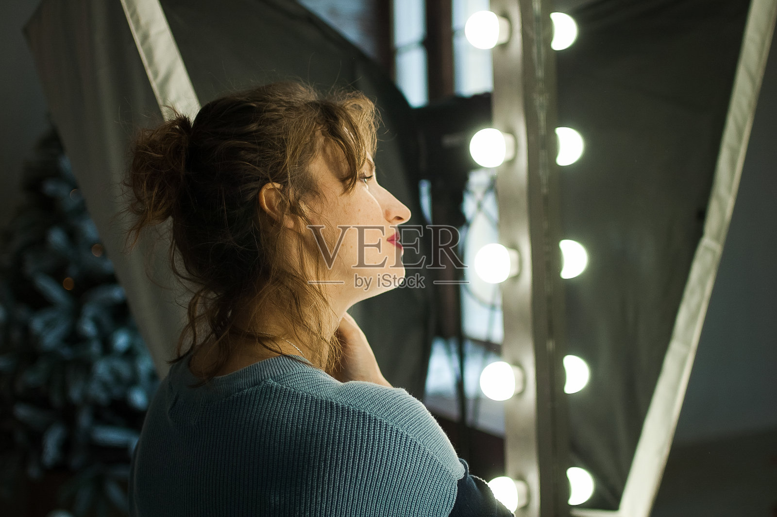 年轻美丽的女人与完美的皮肤在冬季在蓝色毛衣看着镜子在家里的房间圣诞装饰，圣诞树和灯照片摄影图片