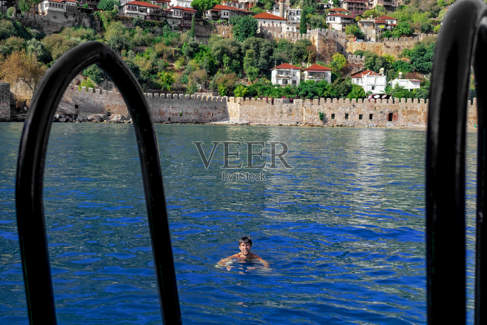 一名男子在蓝色的水中游泳，背景是从甲板上看到的土耳其的阿兰亚古城照片摄影图片