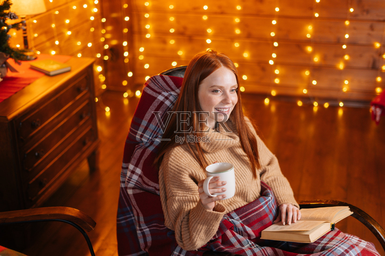 肖像放松的女人拿着杯与热咖啡和读书，而坐在扶手椅照片摄影图片