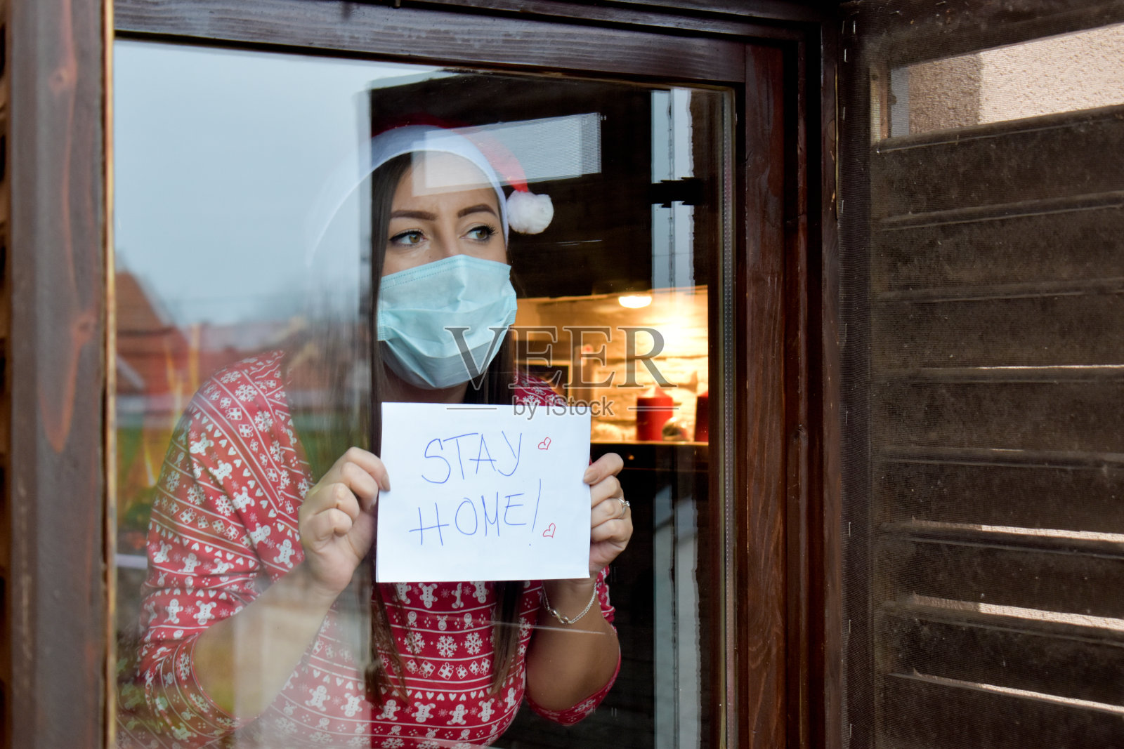 一个戴着面具和圣诞老人帽的女人，站在窗户旁边，举着回家的横幅照片摄影图片