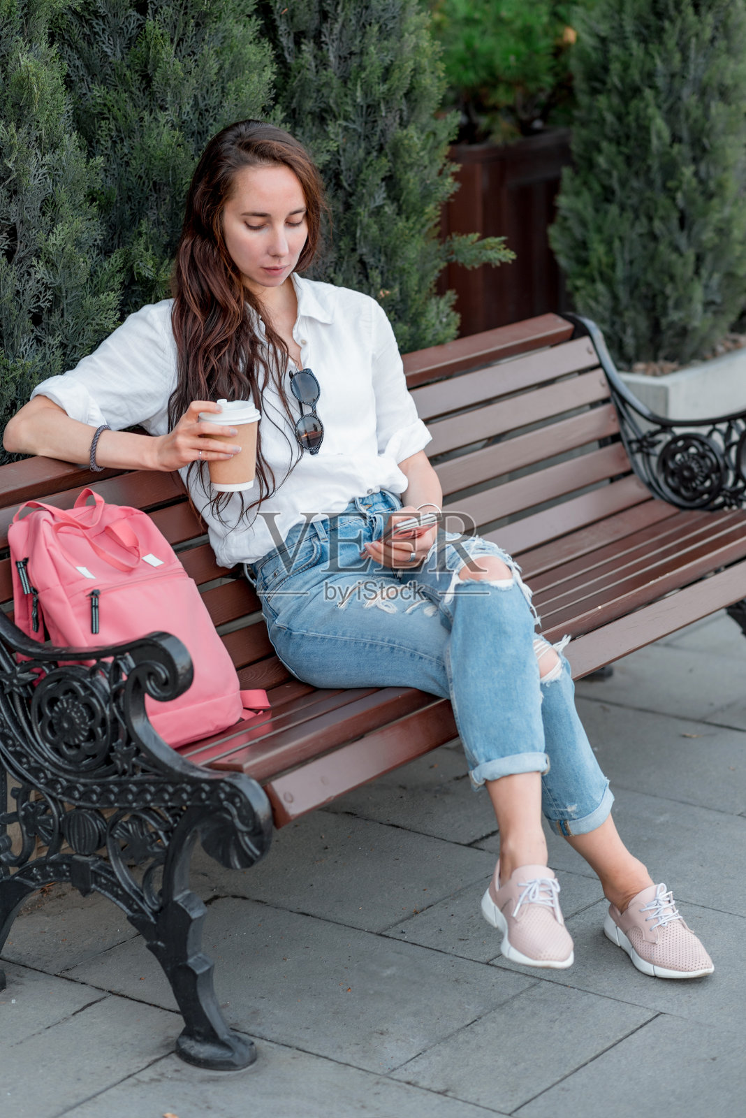 美女坐在夏日城市的长椅上，在手机上在线应用读聊天。白衬衫，牛仔裤，感情一场会一场约，手捧杯咖啡茶。早餐吃零食。长头发的粉色背包。照片摄影图片