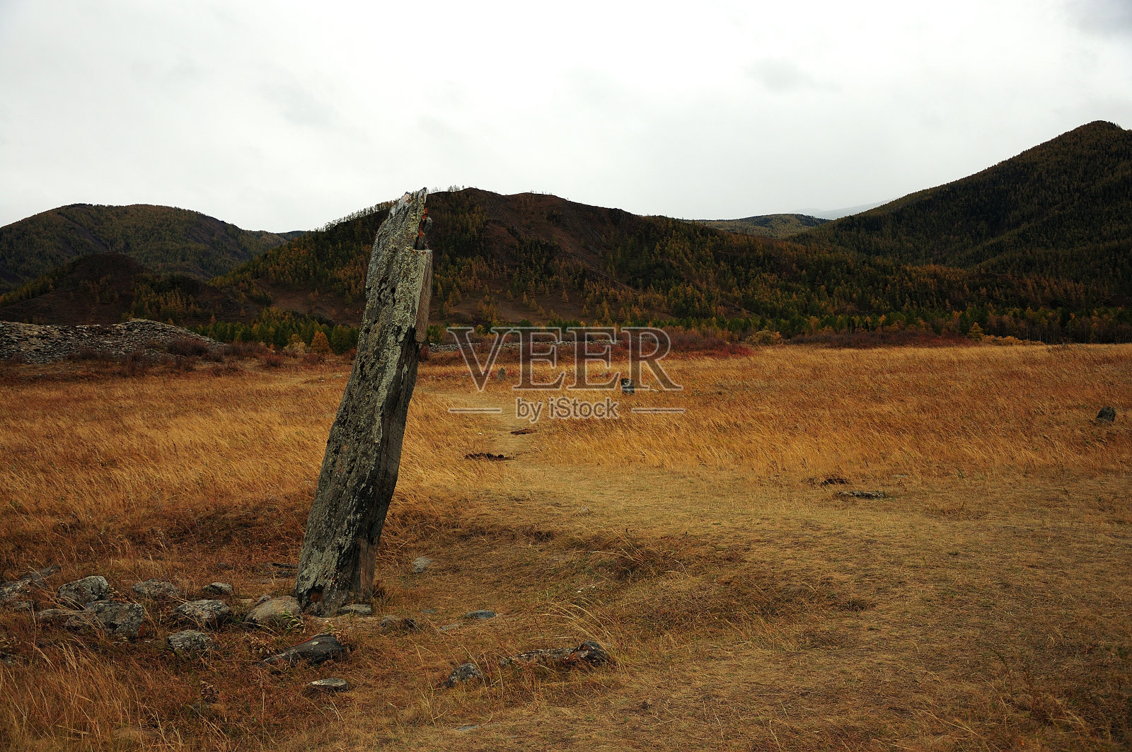 一个古老的石像弯着腰站在秋天的大草原和高山脚下。照片摄影图片