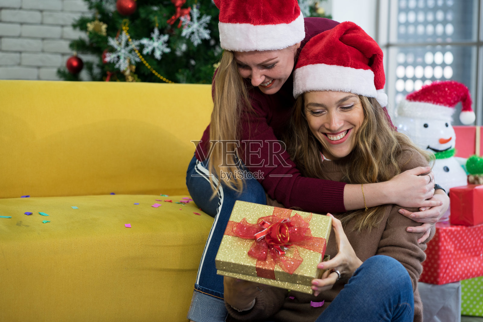 白种女人戴着圣诞老人帽在客厅的黄色沙发上用圣诞树打开礼物盒过节照片摄影图片