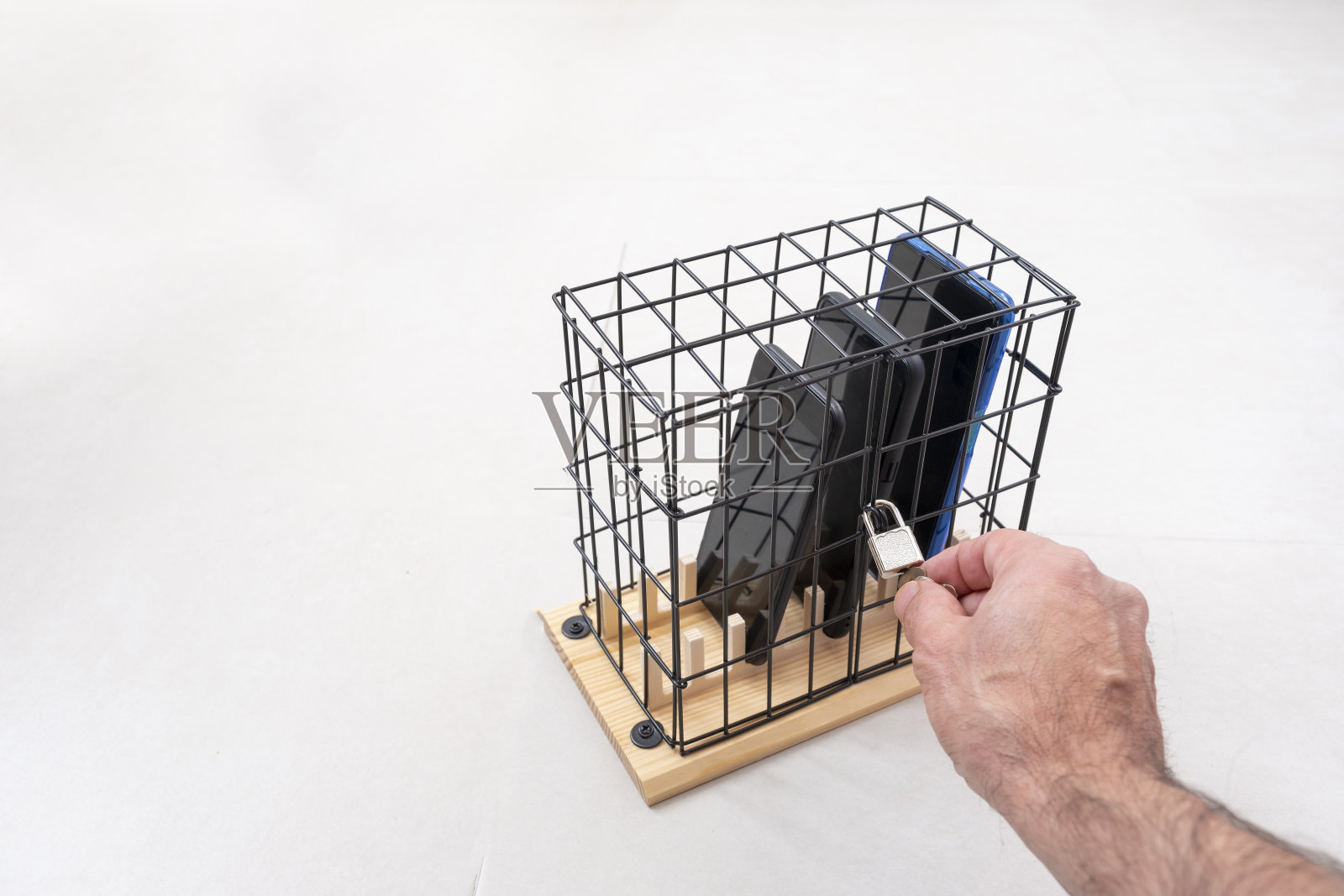 用手打开或关闭一个笼子上的锁，用一个挂锁锁在笼子里的三部手机，社会隔离或手机滥用和社交网络的概念，白色背景，水平照片摄影图片