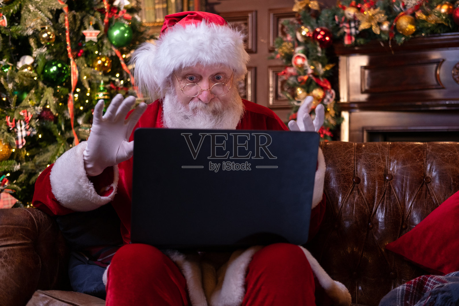 圣诞老人在线祝贺圣诞。圣诞老人用笔记本电脑为远方的圣诞节打电话给坐在圣诞树旁的孩子们。2019年新冠肺炎新年快乐照片摄影图片