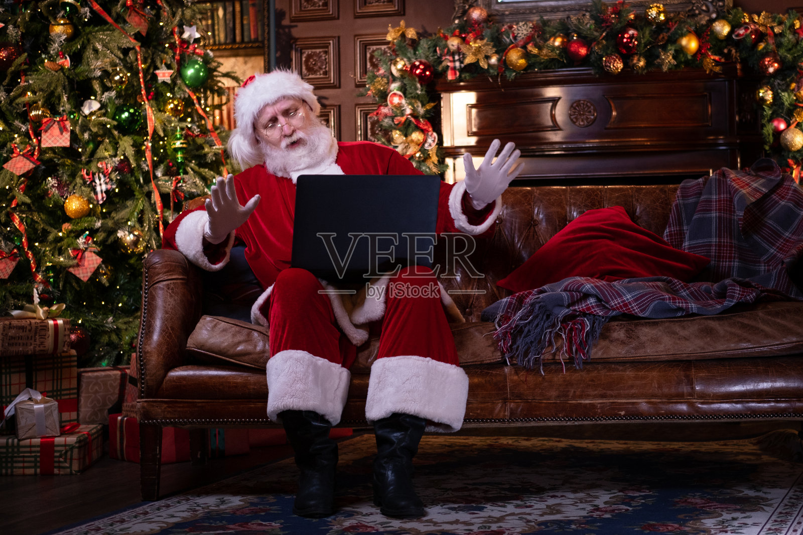 来自圣诞老人的在线祝贺。圣诞老人用笔记本电脑为远方的圣诞节打电话给坐在家里圣诞树旁的孩子。新年快乐照片摄影图片