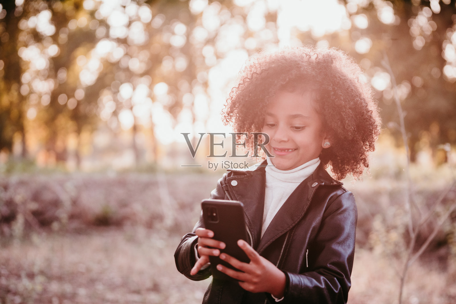 肖像可爱的非洲小女孩使用手机在日落在黄金时间，秋天的季节，美丽的树木背景照片摄影图片