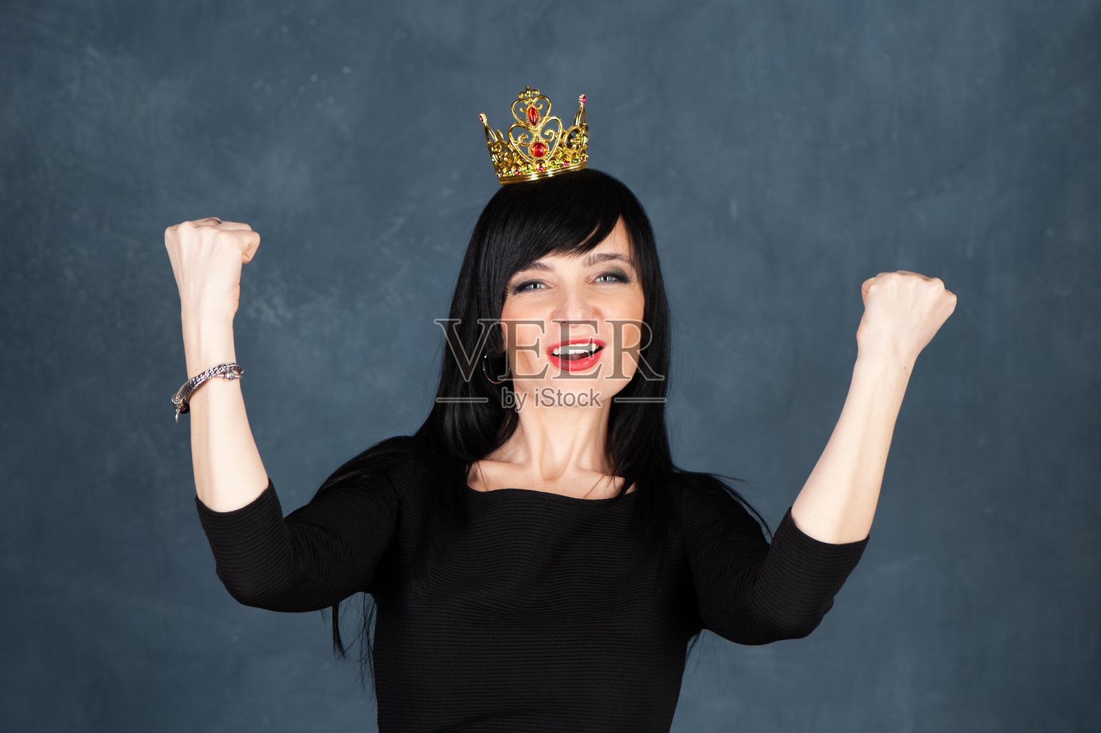 富有表现力和魅力的美丽女孩，深褐色头发，穿着黑色的裙子和皇冠，尖叫着作为幸运和成功的象征。工作室肖像上的单色蓝色，时髦的破旧背景照片摄影图片