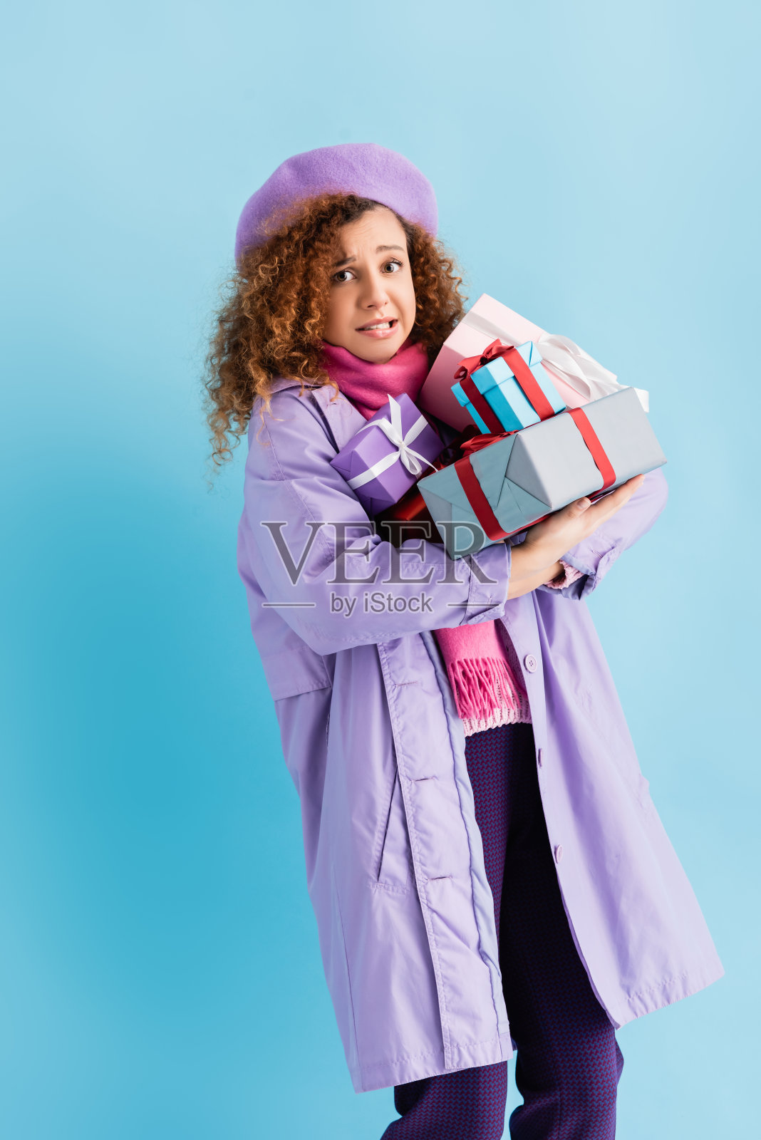 困惑的女人在冬季大衣，贝雷帽和粉红色针织围巾拿着一束圣诞礼物在蓝色照片摄影图片