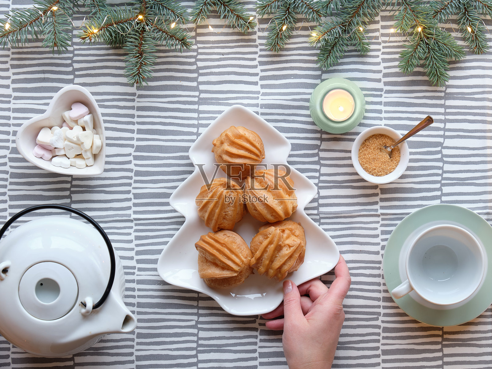 创意圣诞茶几设置。俯视图与泡芙，酝酿蛋糕。手里拿着一个盘子，上面放着圣诞树形状的蛋糕。条纹的纺织桌布上用杉木细枝编成的花环照片摄影图片