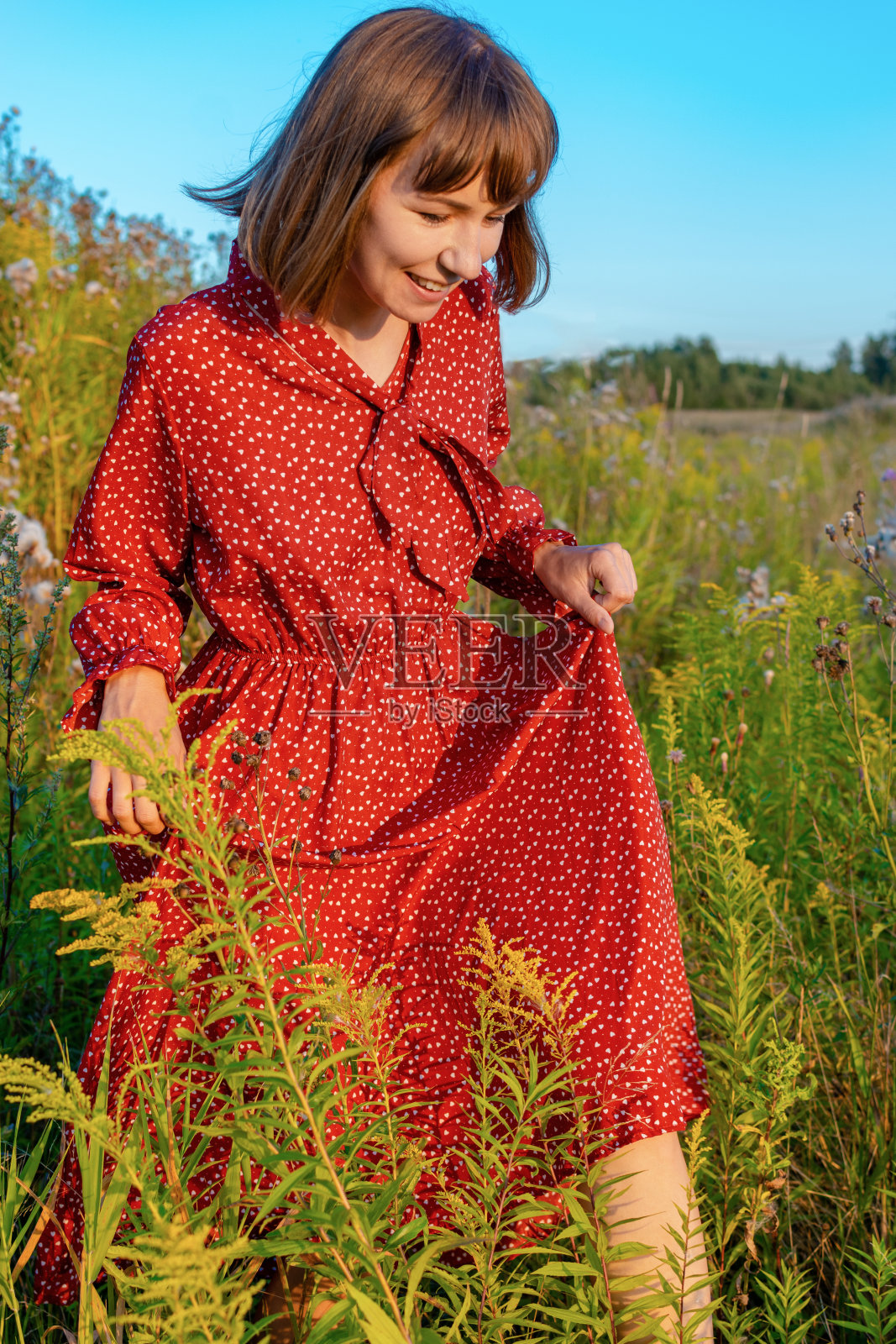 在一个温暖的阳光明媚的日子里，一位穿着红色连衣裙的快乐的年轻女子在田野里散步。照片摄影图片