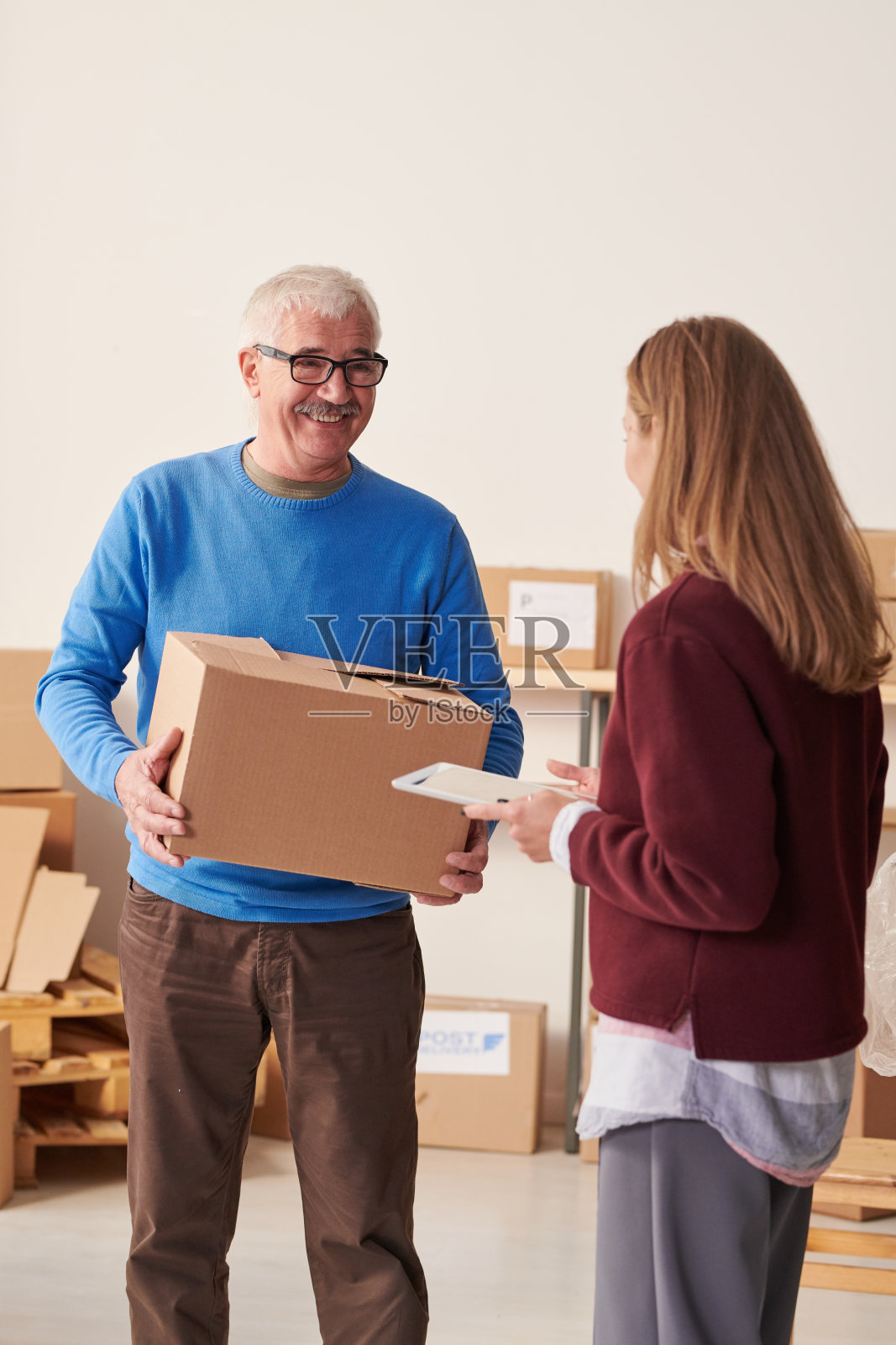 积极英俊的高级仓库员工在蓝色毛衣搬运箱子为调度和谈话经理照片摄影图片