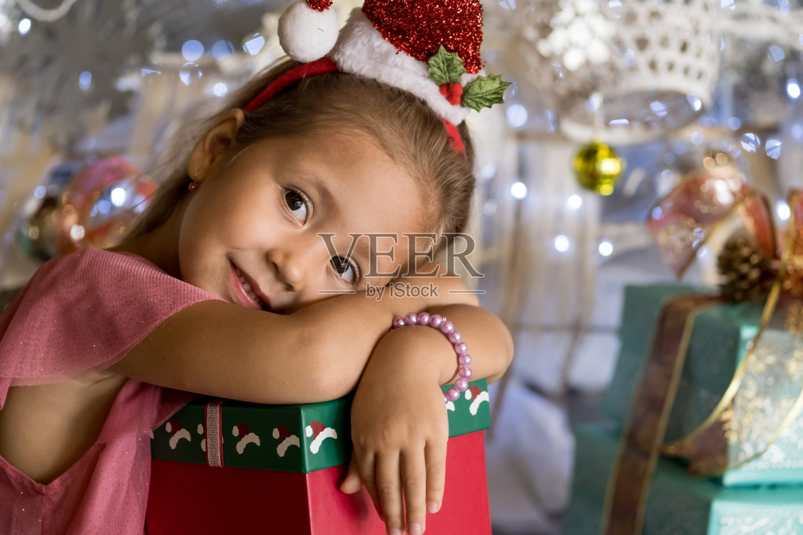 可爱的小女孩坐在圣诞树旁，戴着圣诞帽，拿着礼物盒。圣诞快乐，节日快乐照片摄影图片