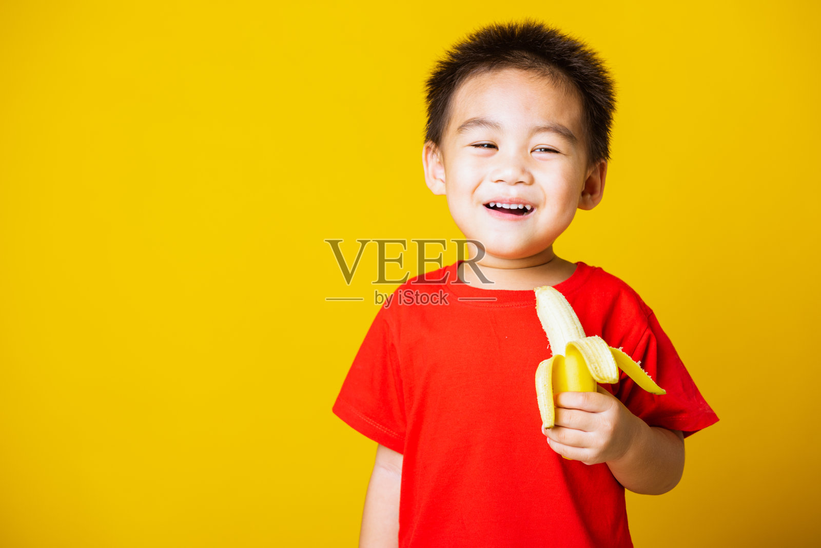 小孩可爱的小男孩迷人的微笑穿着红色的t恤玩着剥了皮的香蕉吃照片摄影图片